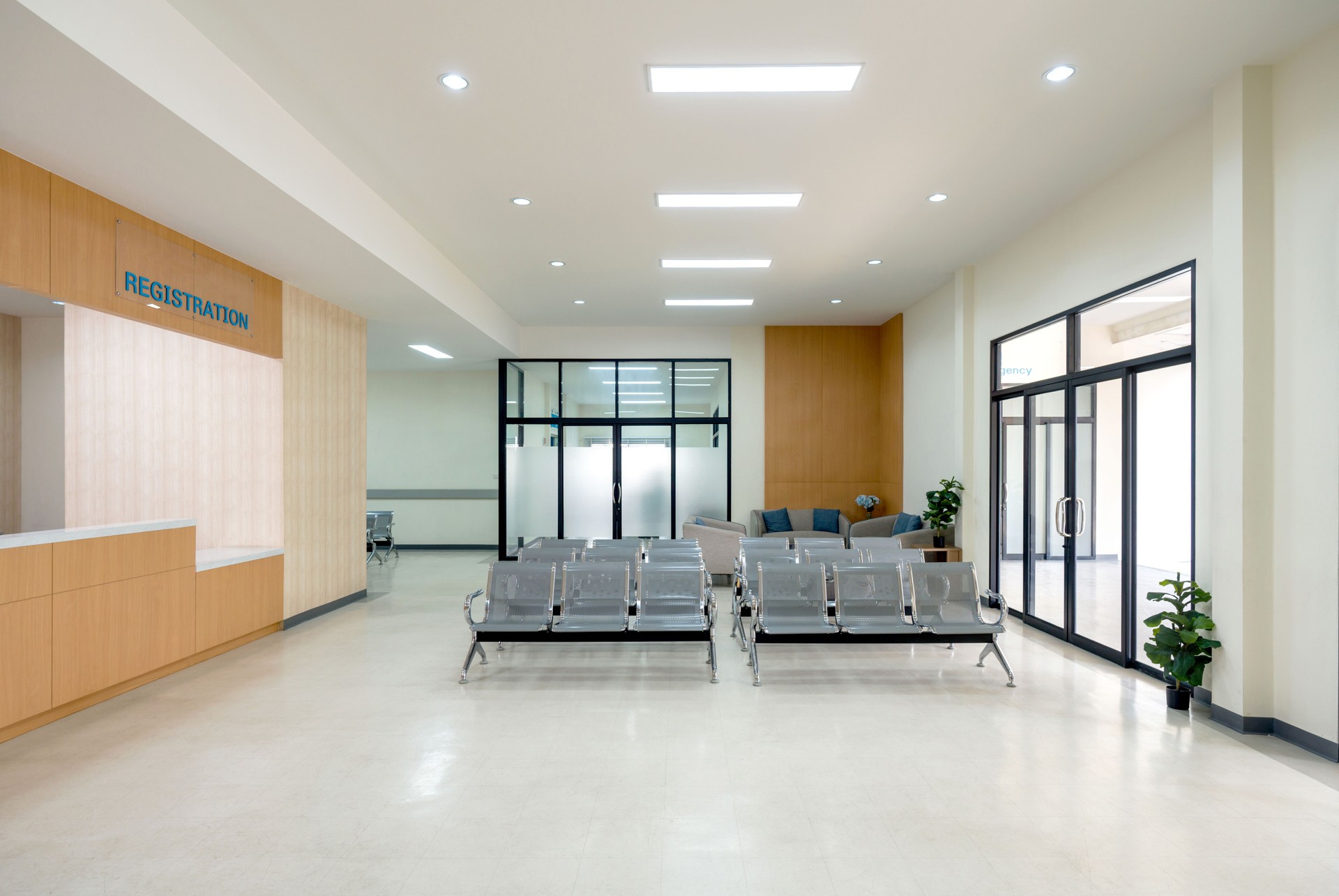 Waiting area in a hospital. Patient or visitor can check in for appointment or service. There are rows of connected grey seating for individual waiting. Glass door on the right lead to emergency area.